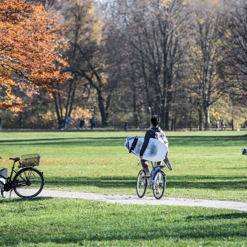 Person riding a bicycle through a park with a surfboard under his arm