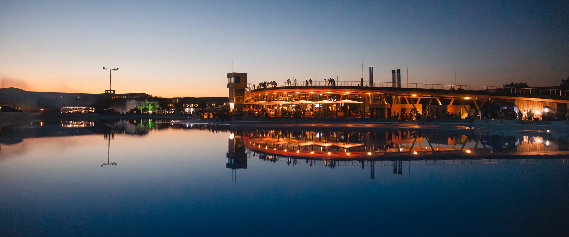 Ob Surfer, Besucher oder Gast im Restaurant oder auf einem Event, die Surftown in M&uuml;nchen bietet Erlebnisse, an die man sich erinnert. Das Bild zeigt eine Aufnahme des Gel&auml;ndes bei D&auml;mmerung, das Wasser der Surfanlage liegt spiegelglatt da und wirft die Reflexion des beleuchteten Geb&auml;udekomplexes mit Restaurant und Walkway &uuml;ber die Rooftop-Terrace. An der Br&uuml;stung sieht man die Silhouetten von Besuchern, die die Aussicht genie&szlig;en.